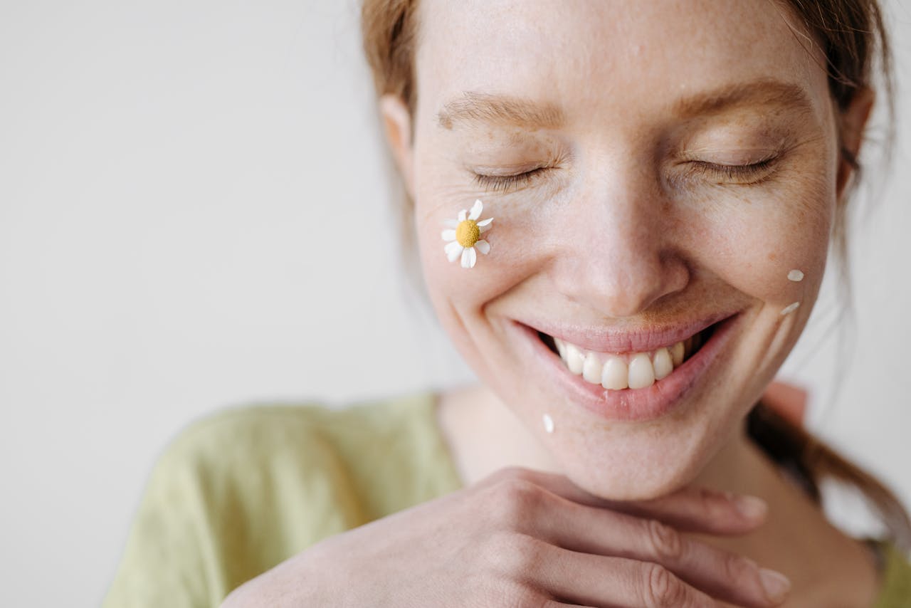 about-img A cheerful woman with a chamomile flower on her cheek, eyes closed in a serene smile.