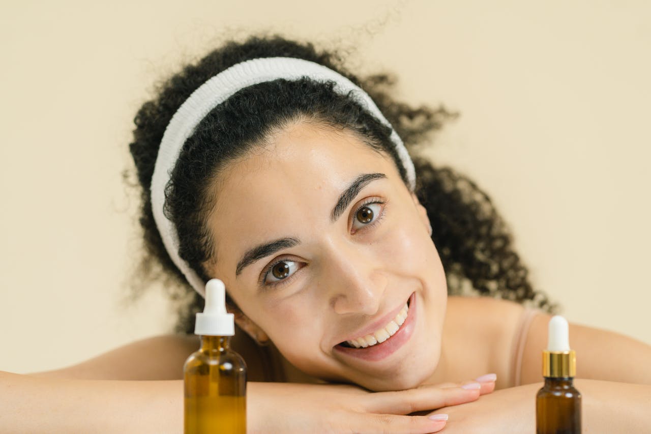 hero-gallery-03 Woman with curly hair smiling, showcasing skincare products, indoors.